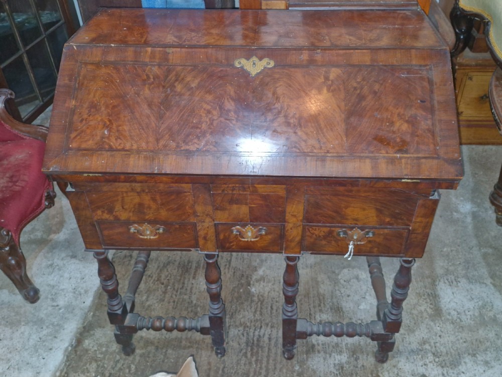A William and Mary style walnut bureau featuring a slanted fall-front writing flap over three lower drawers, supported on turned legs with cross stretchers.