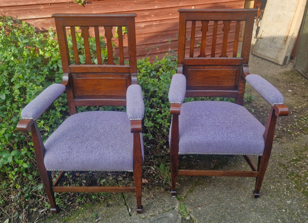 A pair of Edwardian dark oak Arts and Crafts armchairs featuring vertical back splats, upholstered armrests, and purple fabric seats with metal studding.