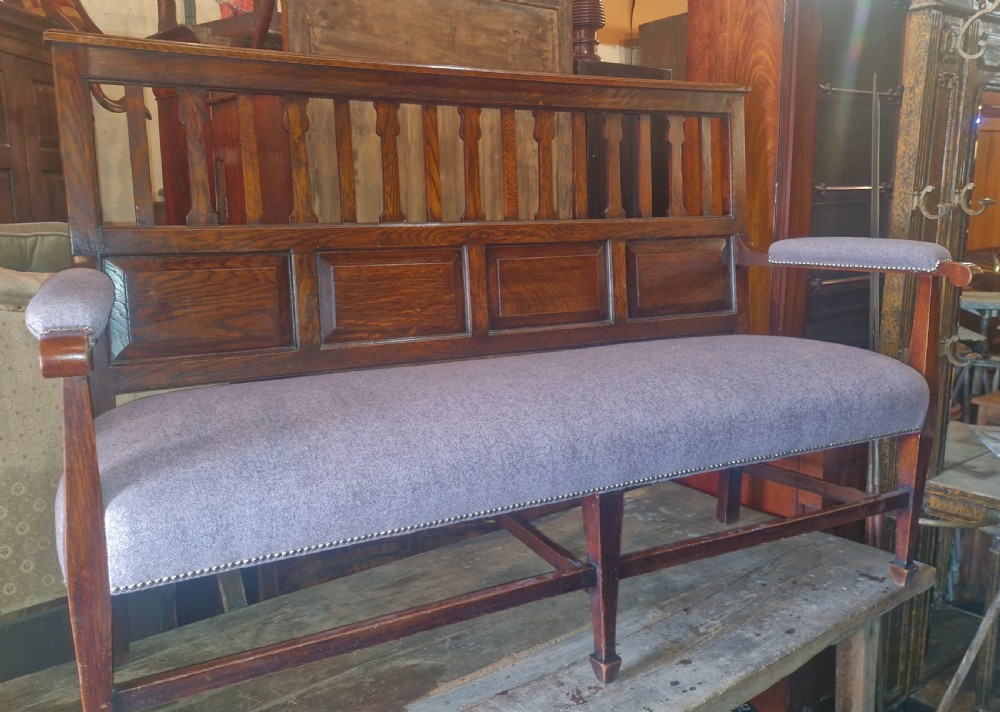 An Edwardian Arts and Crafts dark oak hall bench featuring a slatted and paneled backrest with an upholstered grey seat and padded armrests. View V