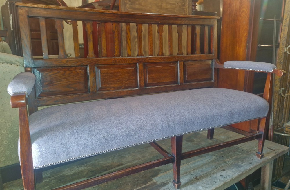 An Edwardian Arts and Crafts dark oak hall bench featuring a slatted and paneled backrest with an upholstered grey seat and padded armrests. View IX