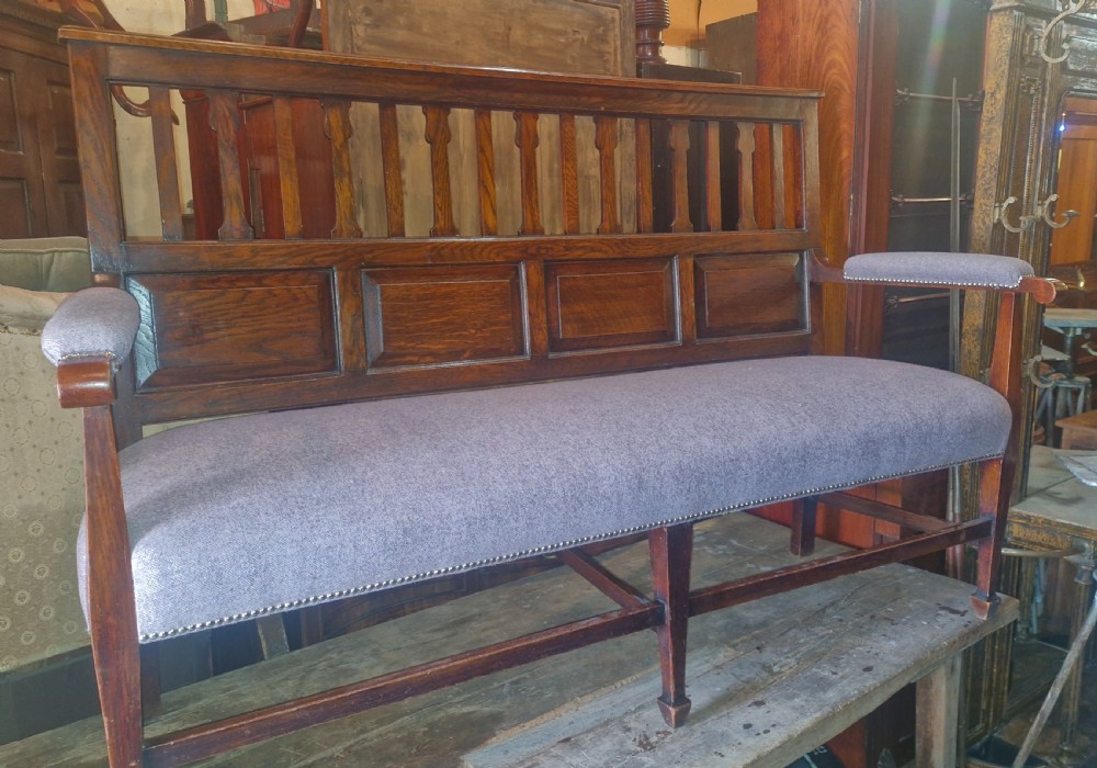 An Edwardian Arts and Crafts dark oak hall bench featuring a slatted and paneled backrest with an upholstered grey seat and padded armrests. View X