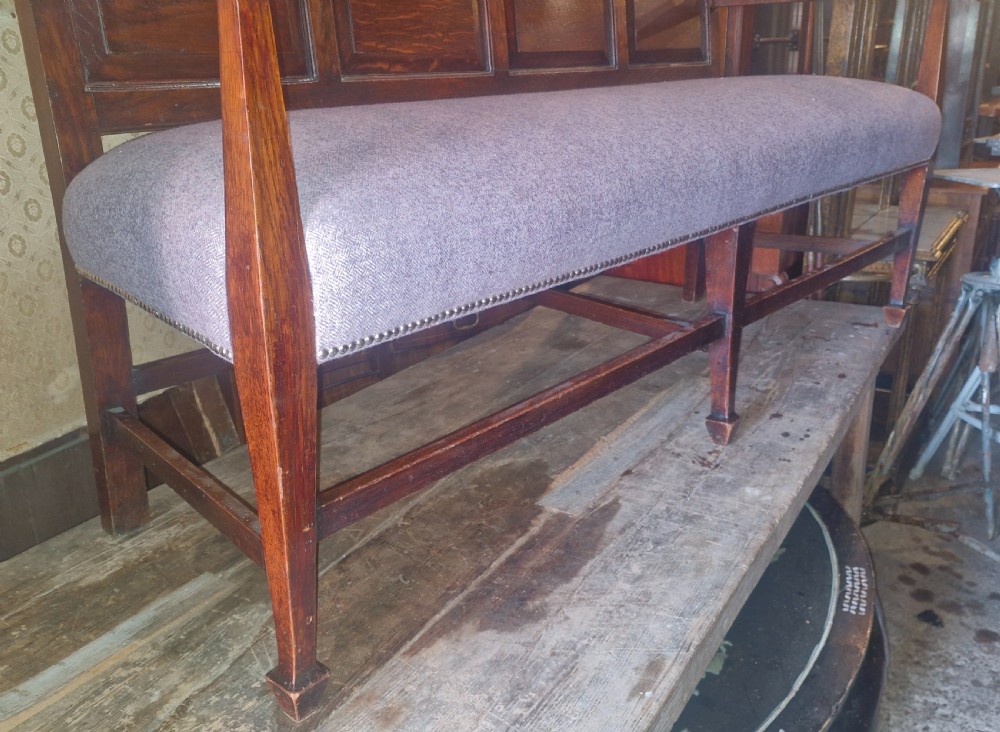 An Edwardian Arts and Crafts dark oak hall bench featuring a slatted and paneled backrest with an upholstered grey seat and padded armrests. View XII