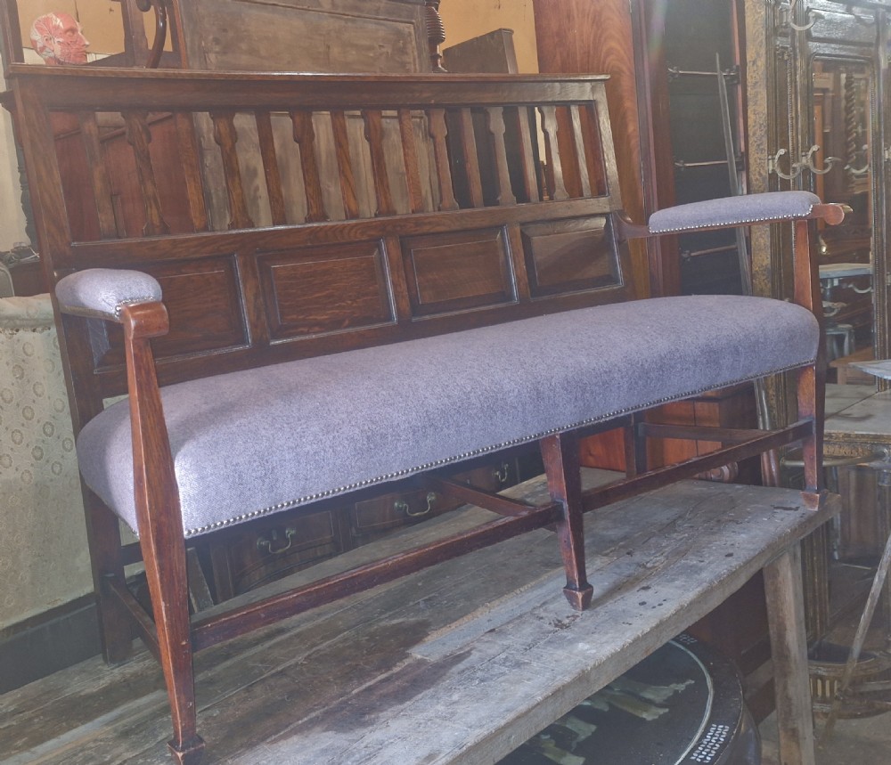 An Edwardian Arts and Crafts dark oak hall bench featuring a slatted and paneled backrest with an upholstered grey seat and padded armrests. View XIII