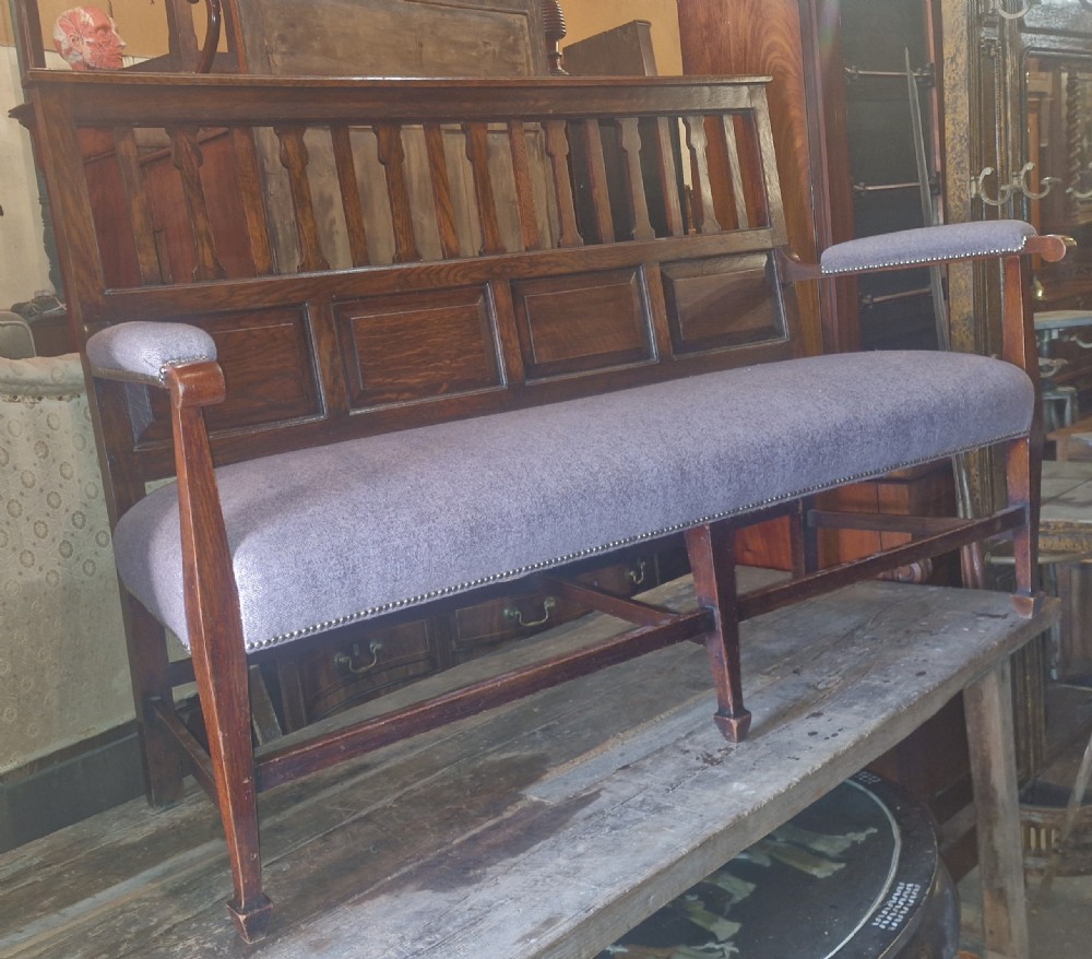An Edwardian Arts and Crafts dark oak hall bench featuring a slatted and paneled backrest with an upholstered grey seat and padded armrests. View XV