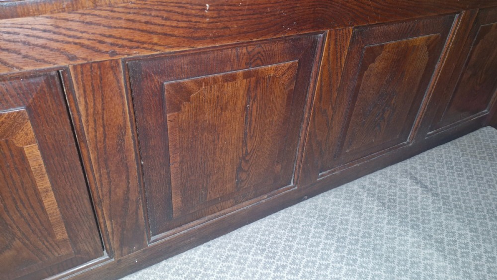 An antique dark oak settle featuring a four-panel backboard, upholstered seat cushion, patterned armrests, and square legs. View V