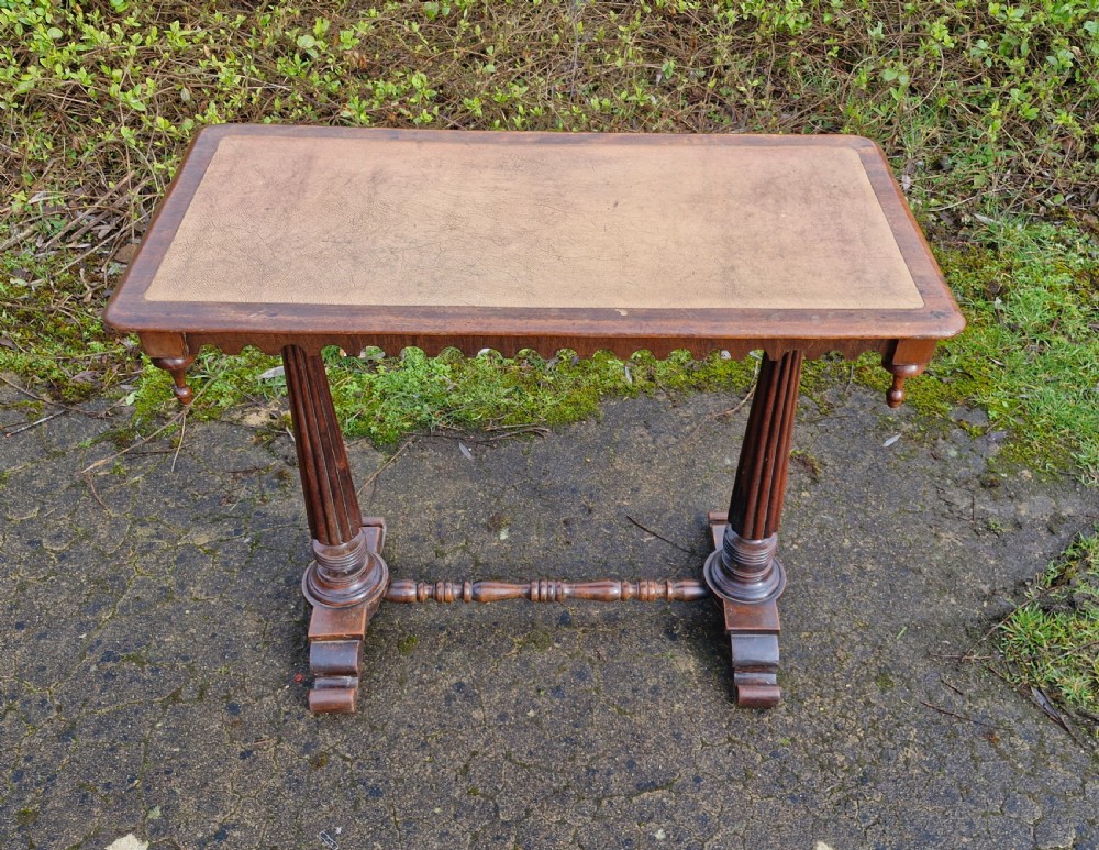 A Victorian mahogany side table featuring a tan leather insert top, scalloped apron, drop finials, and twin fluted column supports joined by a turned stretcher.