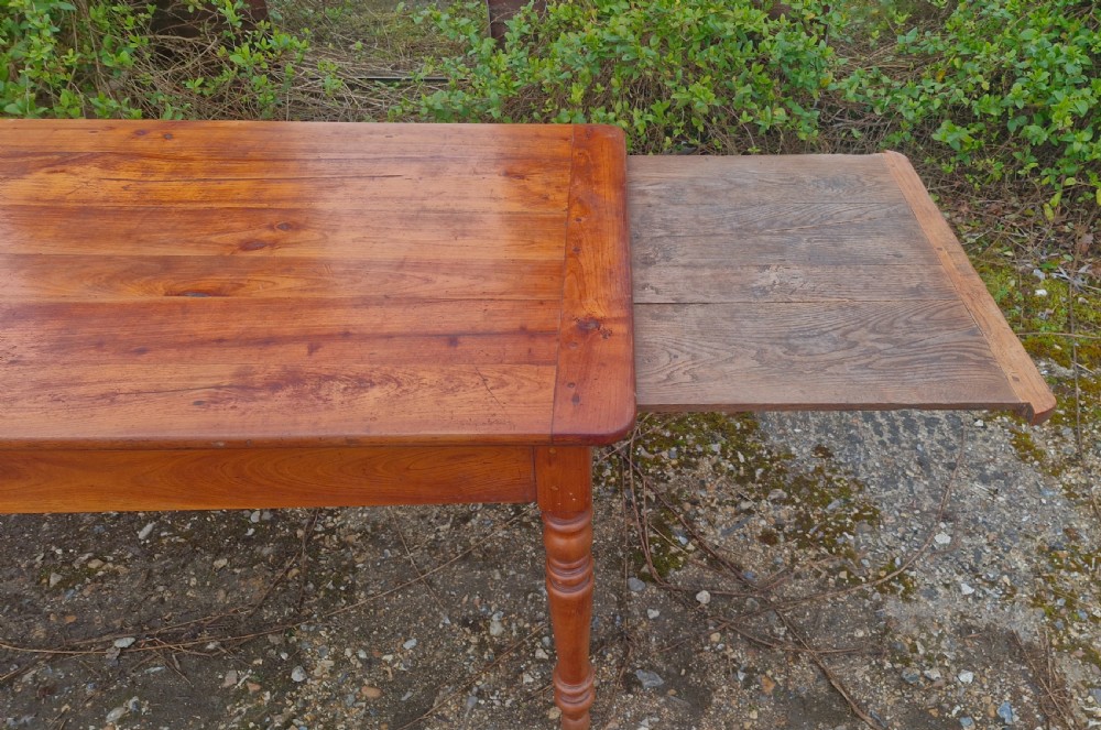 A long rectangular wooden French farmhouse dining table featuring turned legs, an open side drawer, and an extended pull-out bread slide. View VI
