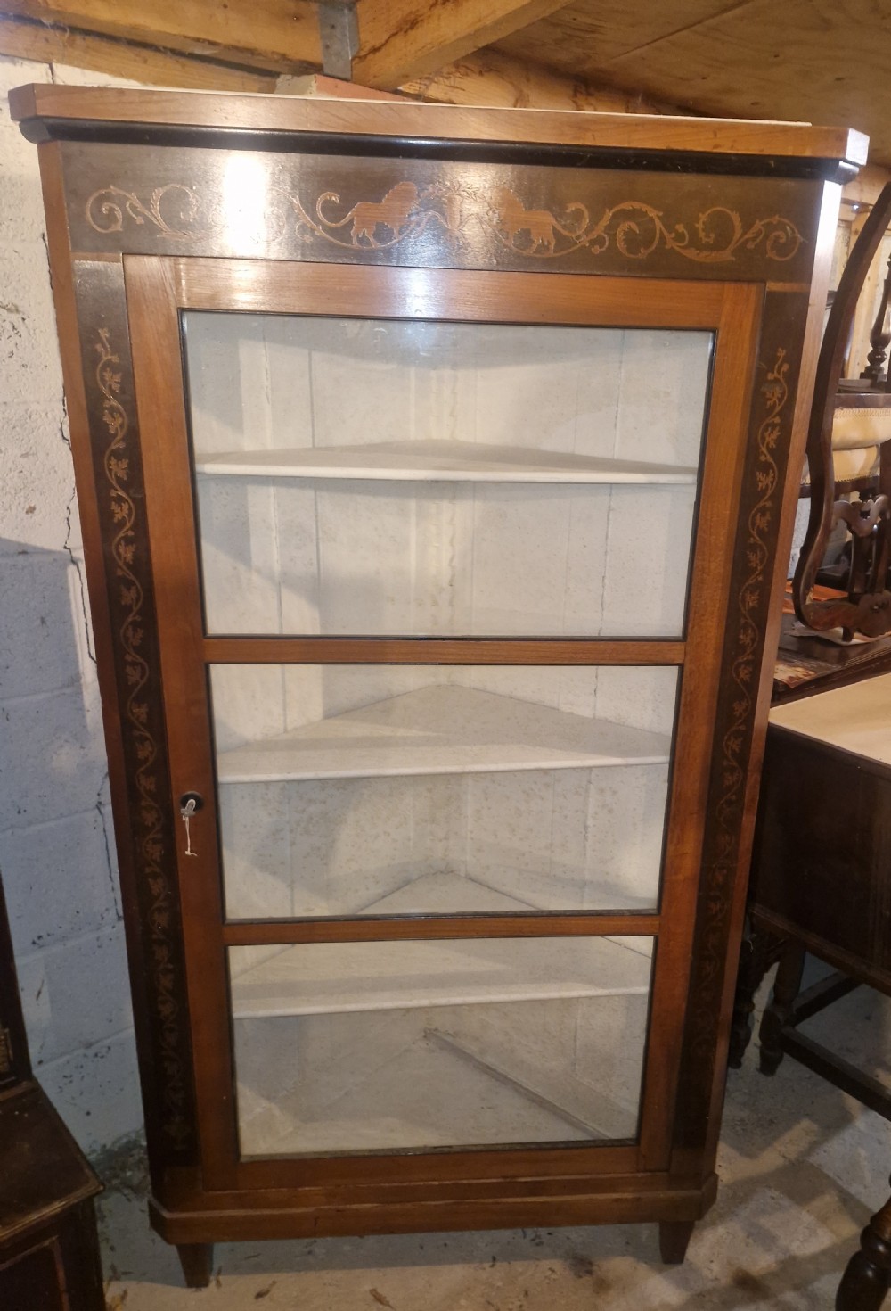 A tall Danish wooden corner cabinet featuring a glazed front door, decorative inlaid scrolling borders, and internal shelves painted off-white.