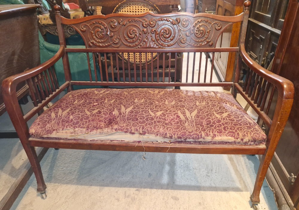 A late Victorian mahogany settee with carved scroll detailing across the top rail, spindle backrest, curved arms, and a patterned red fabric seat on legs with brass castors.