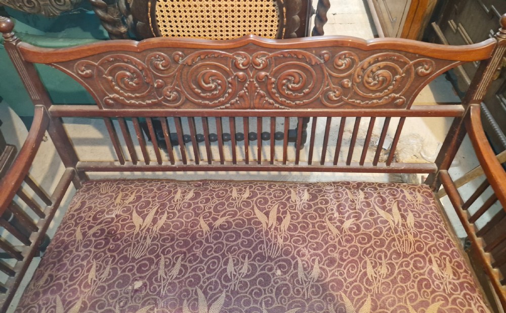 A late Victorian mahogany settee with carved scroll detailing across the top rail, spindle backrest, curved arms, and a patterned red fabric seat on legs with brass castors. View II