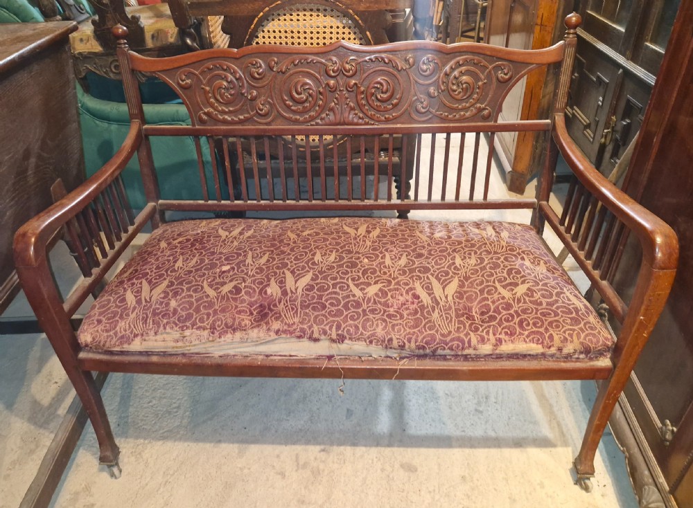 A late Victorian mahogany settee with carved scroll detailing across the top rail, spindle backrest, curved arms, and a patterned red fabric seat on legs with brass castors. View V