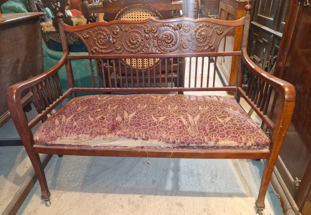 A late Victorian mahogany settee with carved scroll detailing across the top rail, spindle backrest, curved arms, and a patterned red fabric seat on legs with brass castors. View VI