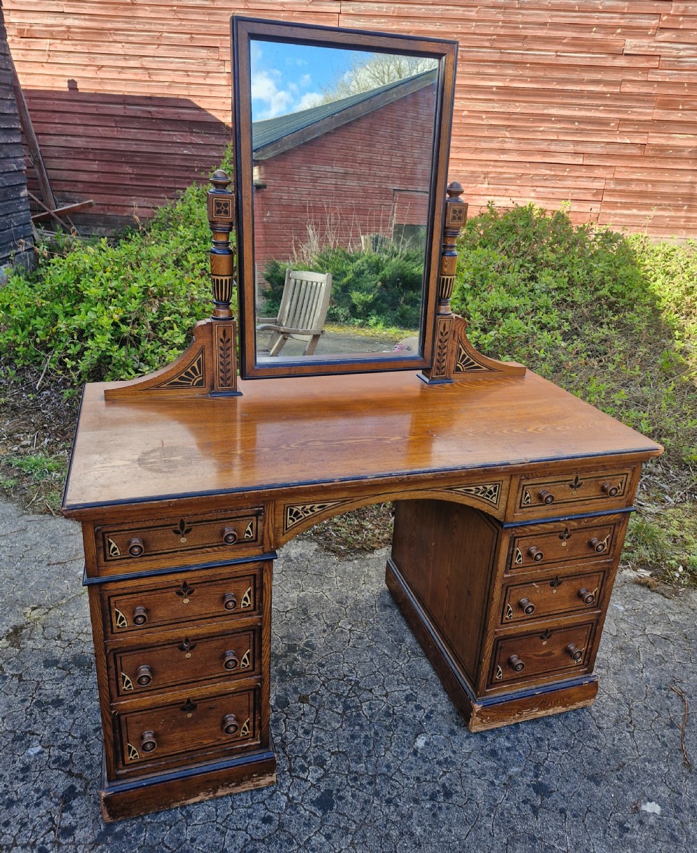 A late Victorian warm brown wood pedestal dressing table featuring ebonised stencilled accents, turned mirror supports, and nine drawers flanking a central kneehole.