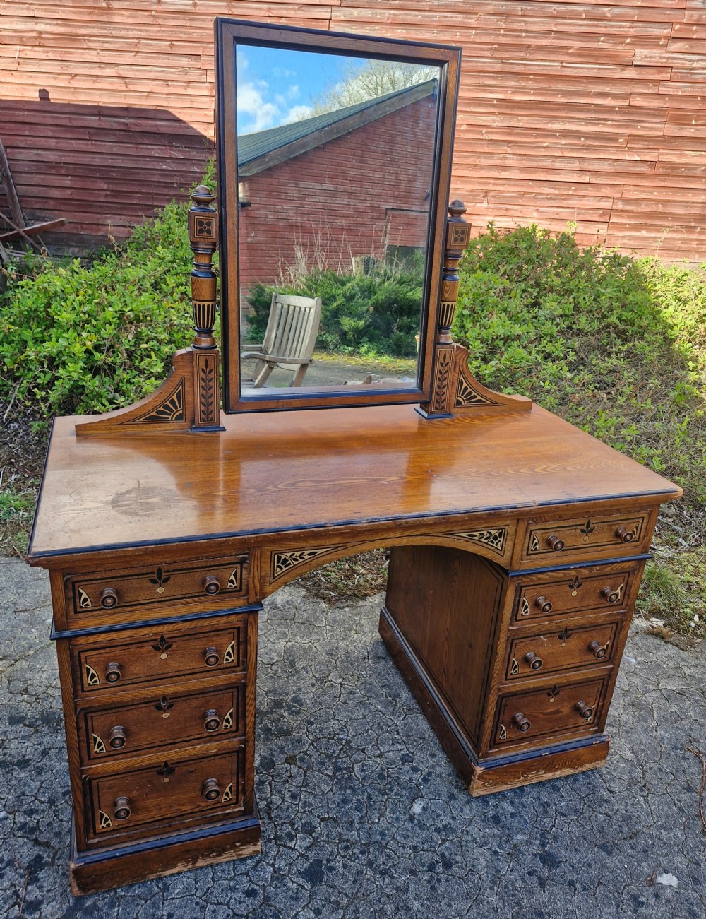 A late Victorian warm brown wood pedestal dressing table featuring ebonised stencilled accents, turned mirror supports, and nine drawers flanking a central kneehole. View X