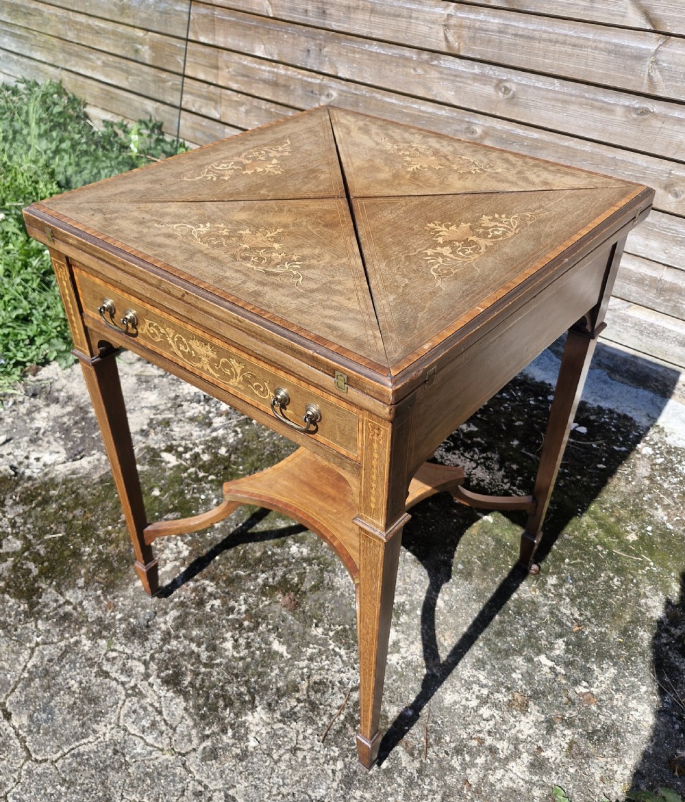 An Edwardian square mahogany envelope games table featuring foliate marquetry inlay, a single drawer with brass drop handles, a lower undertier, and tapered legs.