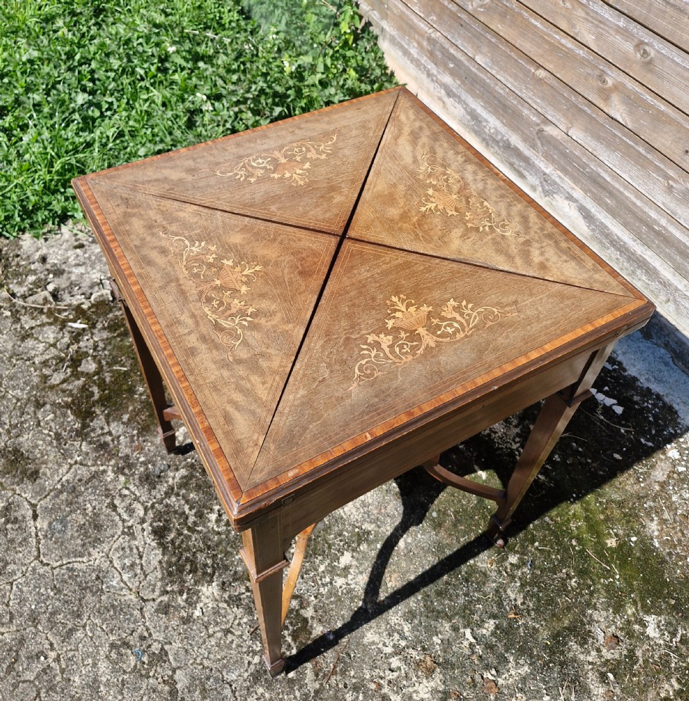 An Edwardian square mahogany envelope games table featuring foliate marquetry inlay, a single drawer with brass drop handles, a lower undertier, and tapered legs. View V