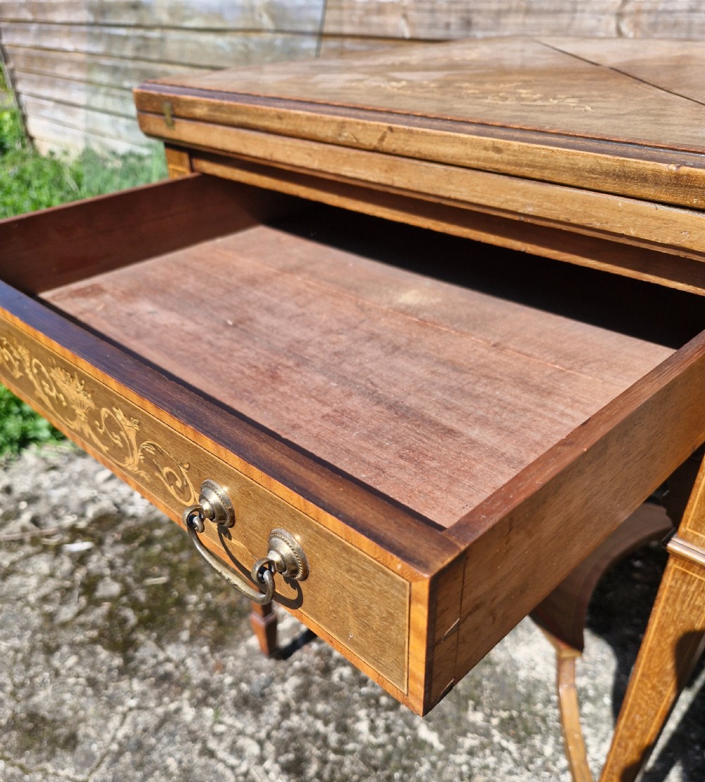 An Edwardian square mahogany envelope games table featuring foliate marquetry inlay, a single drawer with brass drop handles, a lower undertier, and tapered legs. View VI
