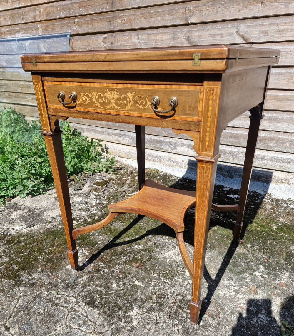 An Edwardian square mahogany envelope games table featuring foliate marquetry inlay, a single drawer with brass drop handles, a lower undertier, and tapered legs. View VII