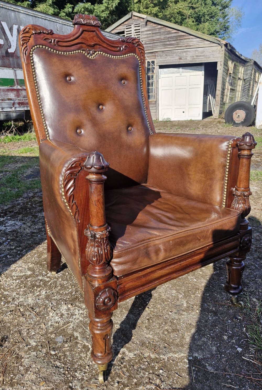 A Regency style mahogany library armchair upholstered in button-tufted brown leather with carved detailing and brass castors.