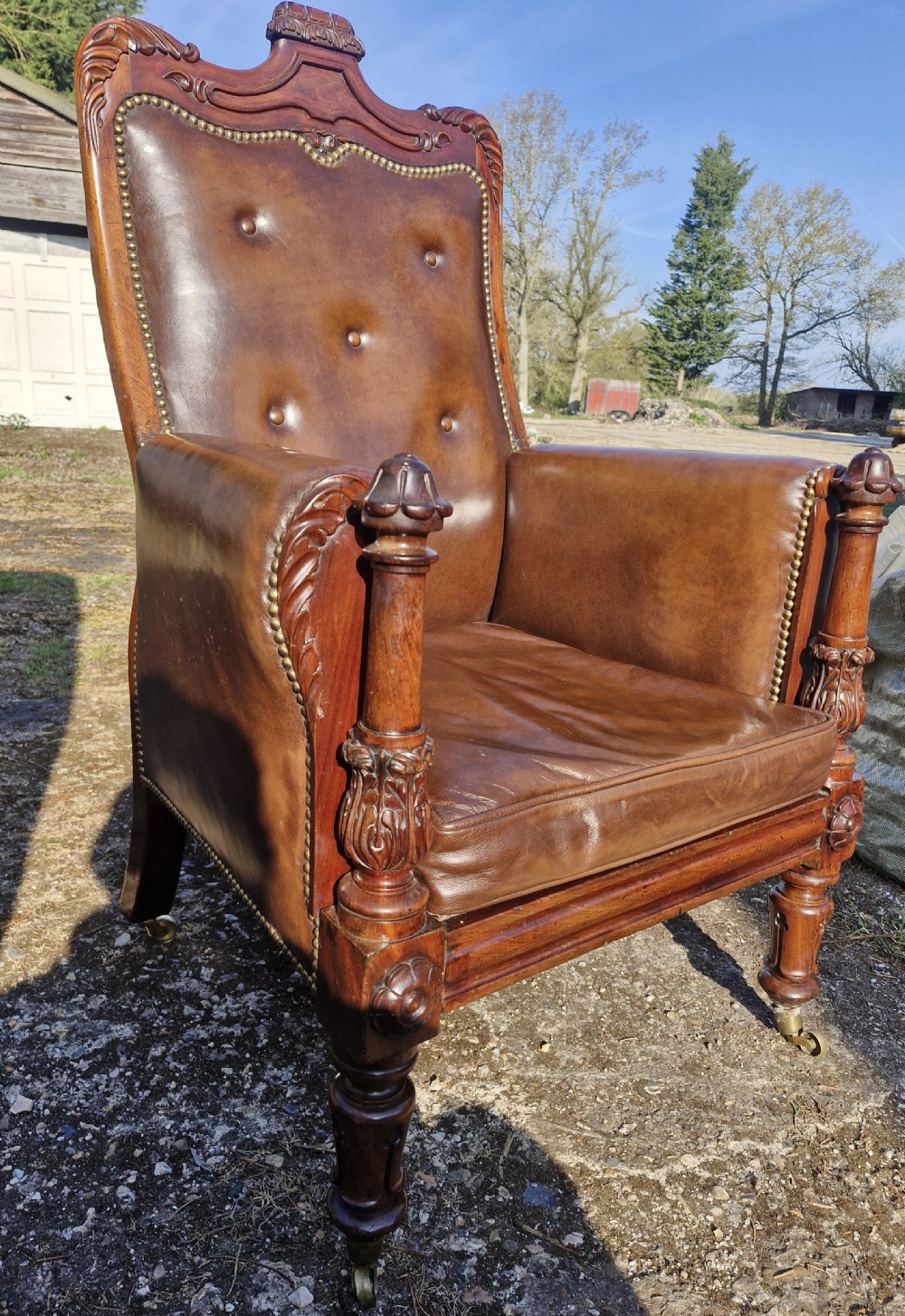 A Regency style mahogany library armchair upholstered in button-tufted brown leather with carved detailing and brass castors. View VI