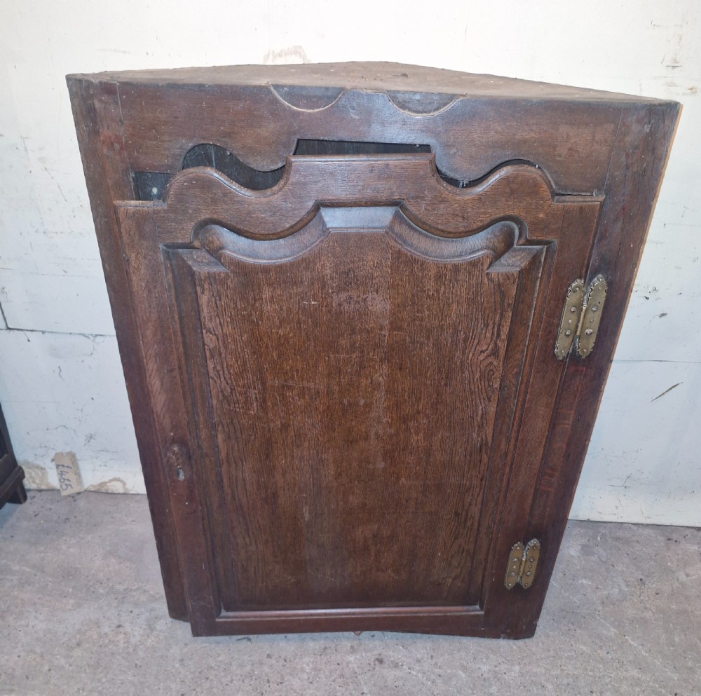 An antique dark oak French corner cupboard featuring a single carved panelled door with brass hinges and a shaped top outline. View VI