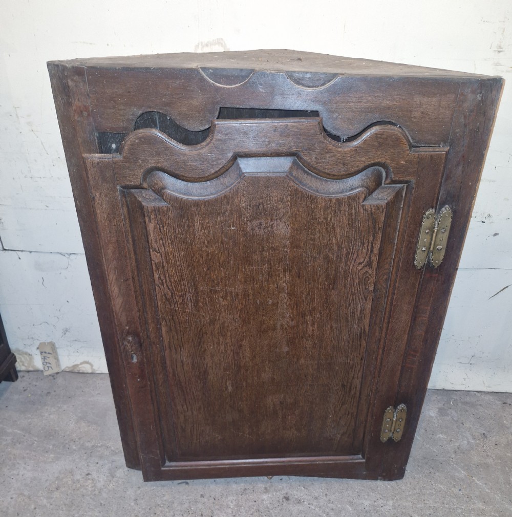 An antique dark oak French corner cupboard featuring a single carved panelled door with brass hinges and a shaped top outline.