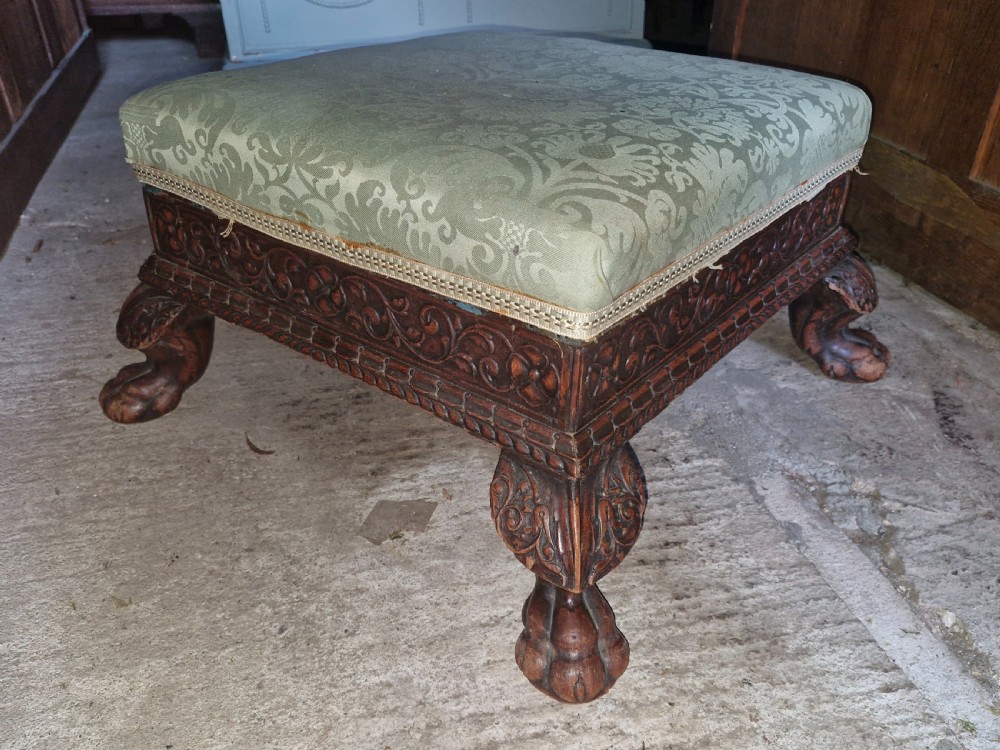 A square Anglo-Indian footstool featuring a green damask upholstered top above a deeply carved dark hardwood frame with claw feet.