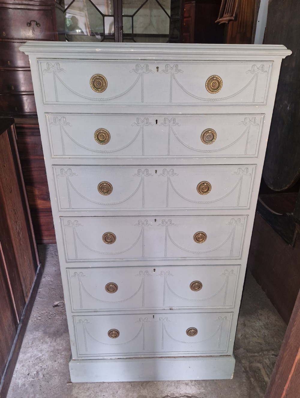 An Edwardian light grey painted pine Wellington chest featuring six drawers decorated with carved ribbon swag details and brass ring handles.