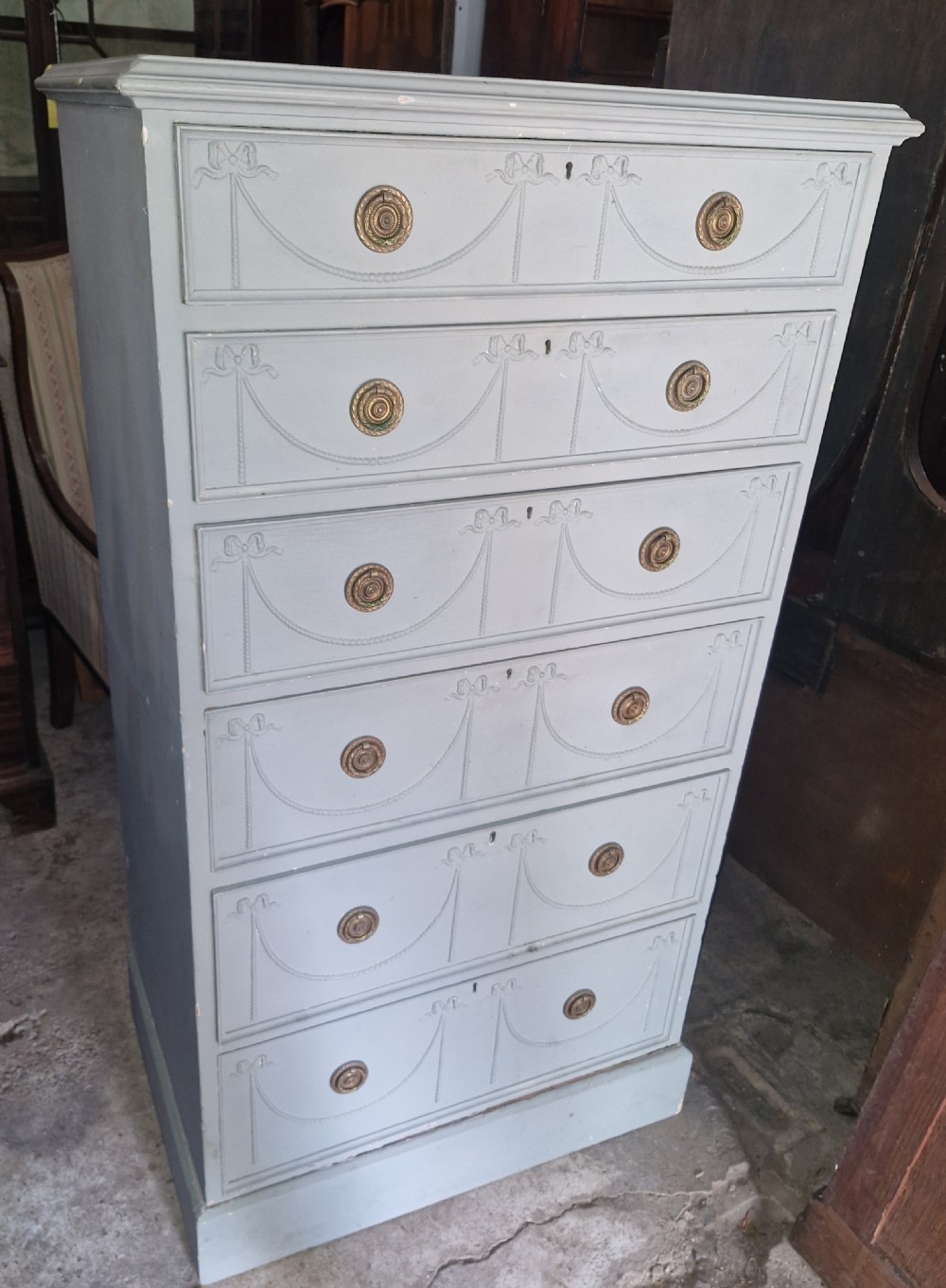An Edwardian light grey painted pine Wellington chest featuring six drawers decorated with carved ribbon swag details and brass ring handles. View V