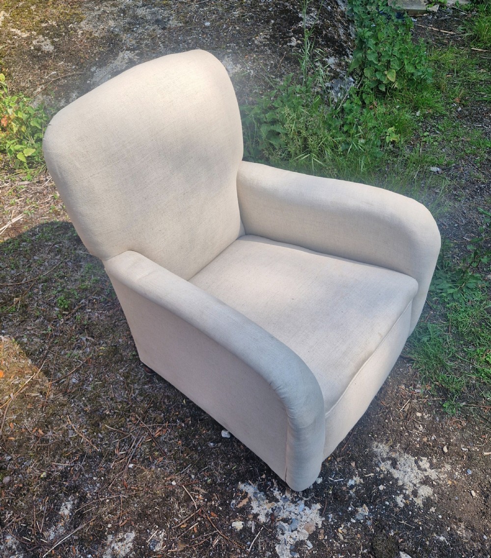 A fully upholstered early 20th century childs armchair featuring neutral beige fabric, rolled arms, and a curved backrest.