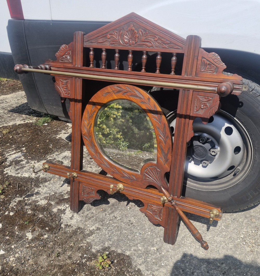 A late Victorian carved dark wood wall-mounted hat and coat rack featuring a central oval mirror, turned spindles, a brass rail, and brass hooks.
