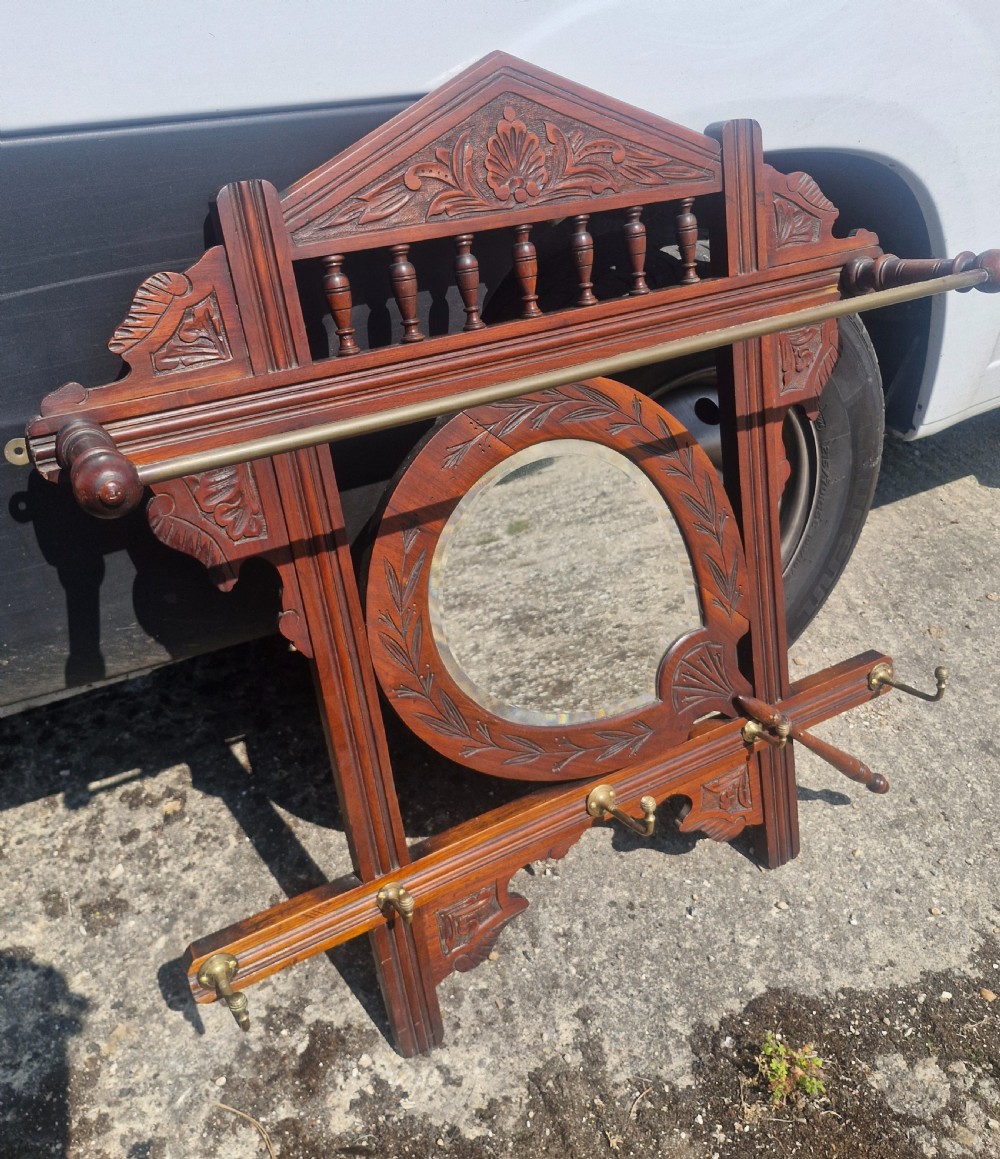 A late Victorian carved dark wood wall-mounted hat and coat rack featuring a central oval mirror, turned spindles, a brass rail, and brass hooks. View II