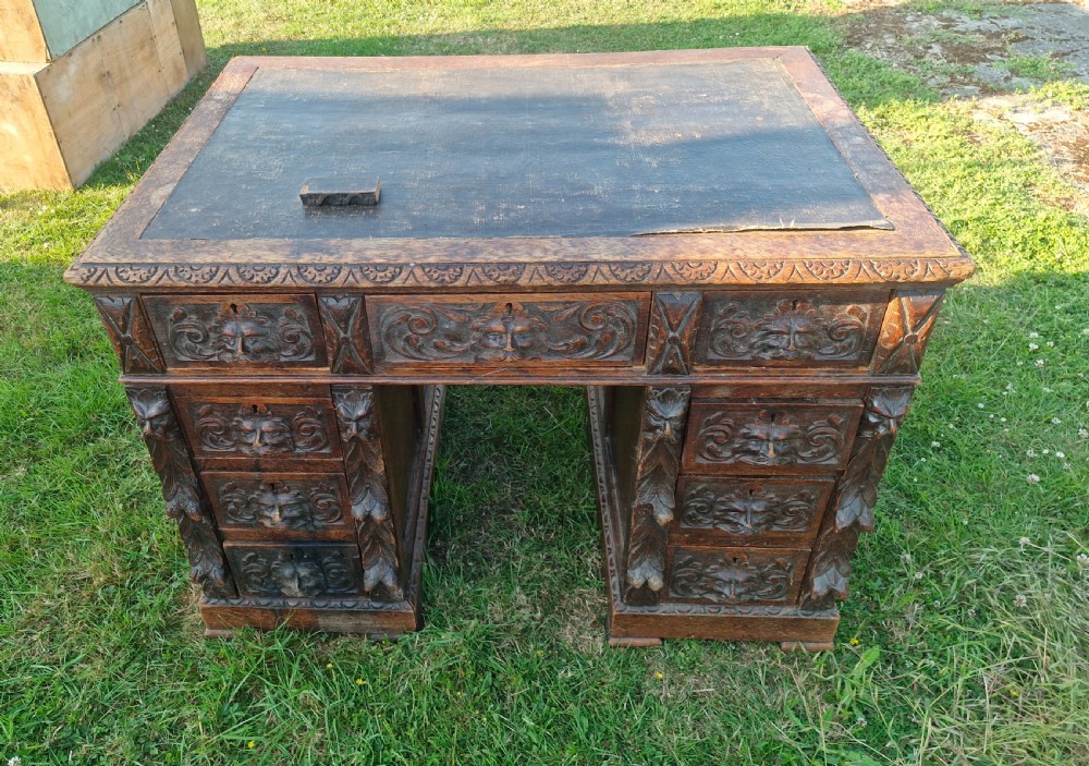 A carved dark oak twin-pedestal desk featuring relief mask drawer handles, foliate side columns, and an inset dark leather writing surface. View V