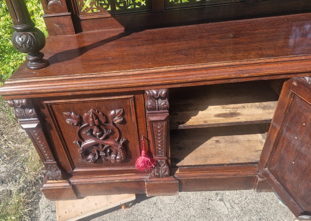An ornate dark oak Victorian buffet featuring relief-carved cabinet doors, turned support columns, openwork fretwork back panels, and an arched top pediment with carved swag detailing. View II