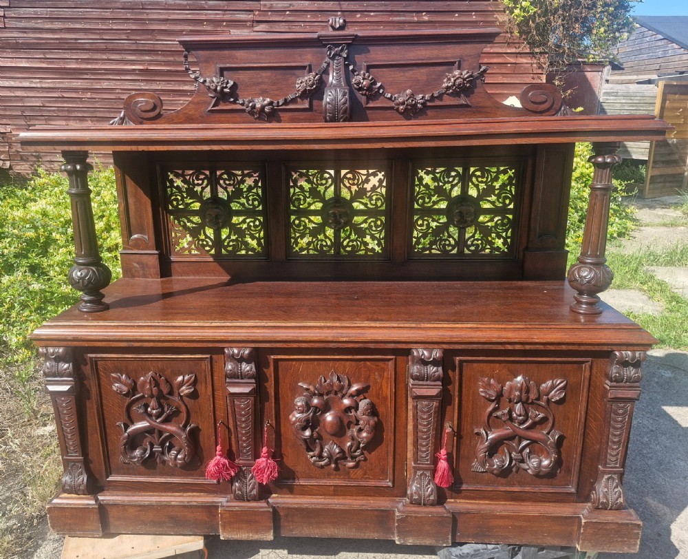 An ornate dark oak Victorian buffet featuring relief-carved cabinet doors, turned support columns, openwork fretwork back panels, and an arched top pediment with carved swag detailing. View III