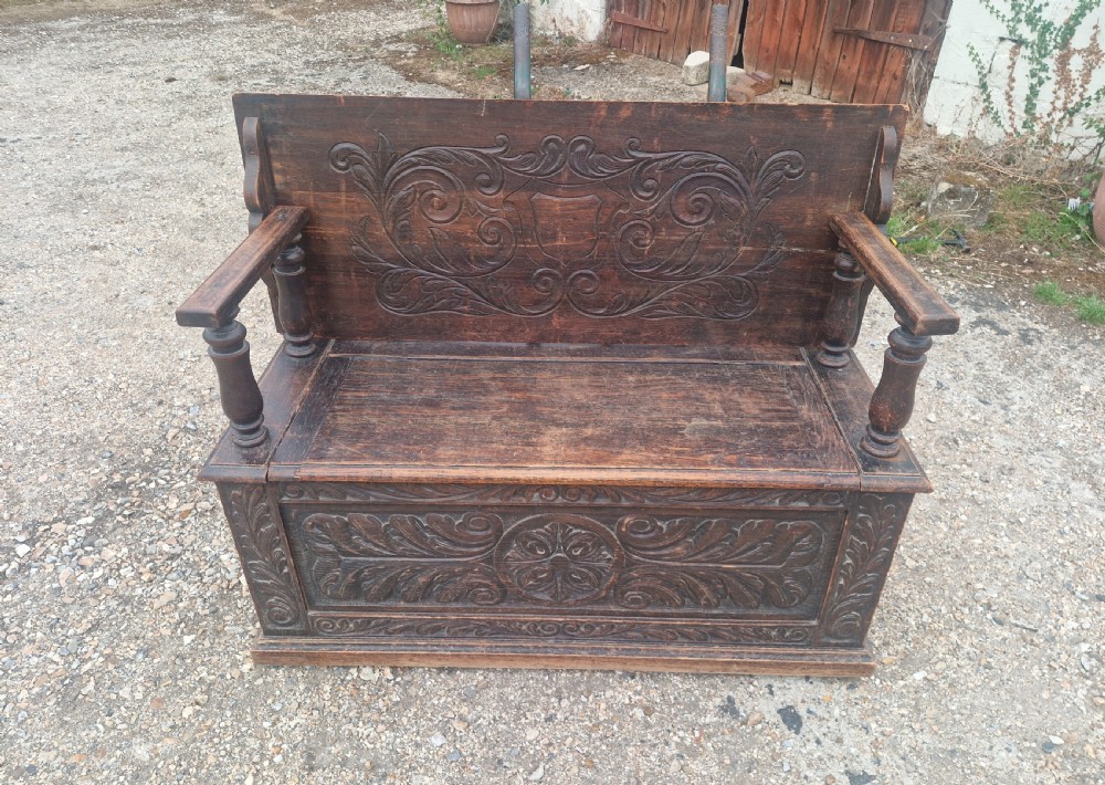 Late Victorian dark oak monks hall seat featuring intricate carved foliate panels, turned arm supports, and an integrated storage chest base.