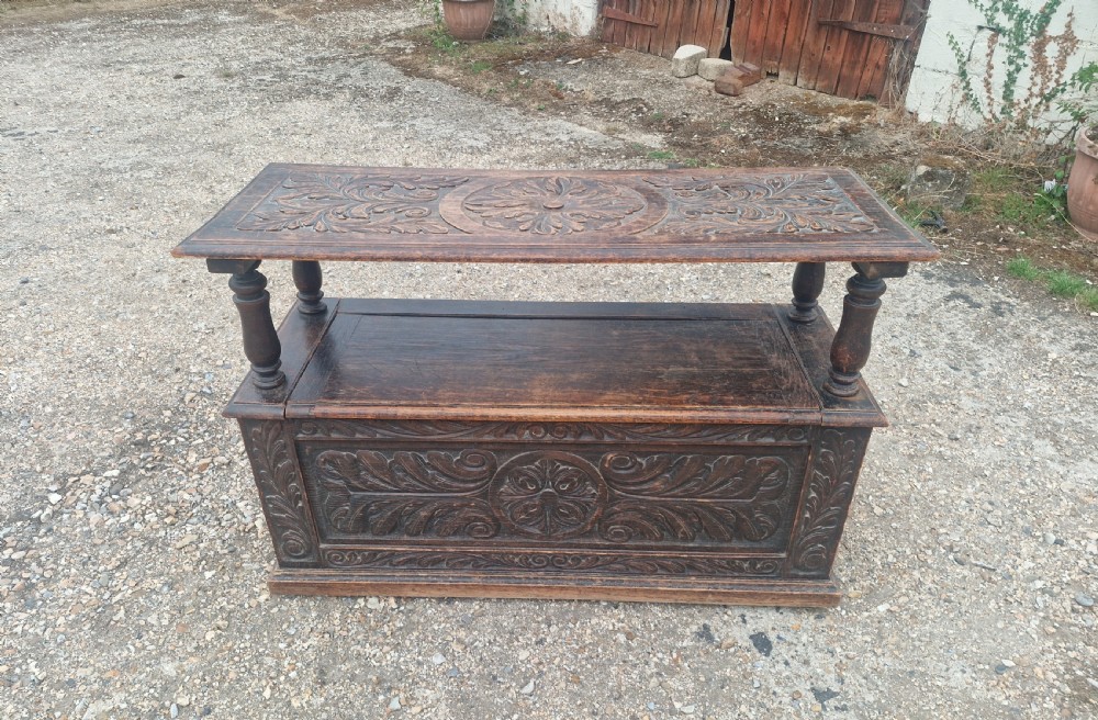 Late Victorian dark oak monks hall seat featuring intricate carved foliate panels, turned arm supports, and an integrated storage chest base. View II