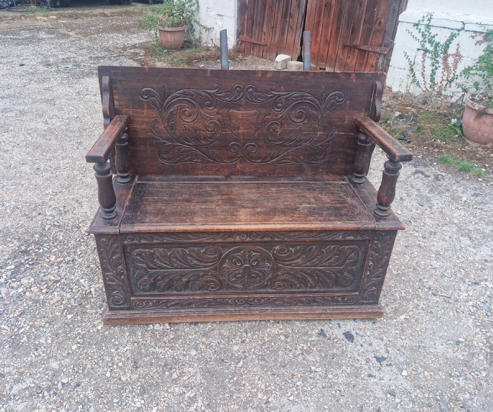 Late Victorian dark oak monks hall seat featuring intricate carved foliate panels, turned arm supports, and an integrated storage chest base. View III