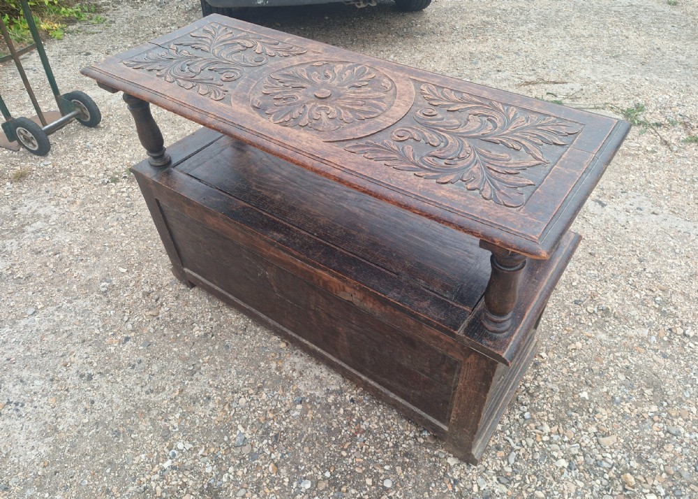 Late Victorian dark oak monks hall seat featuring intricate carved foliate panels, turned arm supports, and an integrated storage chest base. View V