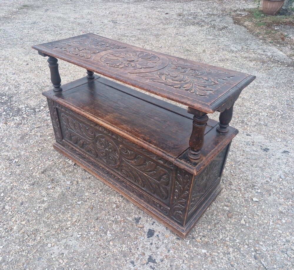 Late Victorian dark oak monks hall seat featuring intricate carved foliate panels, turned arm supports, and an integrated storage chest base. View VI