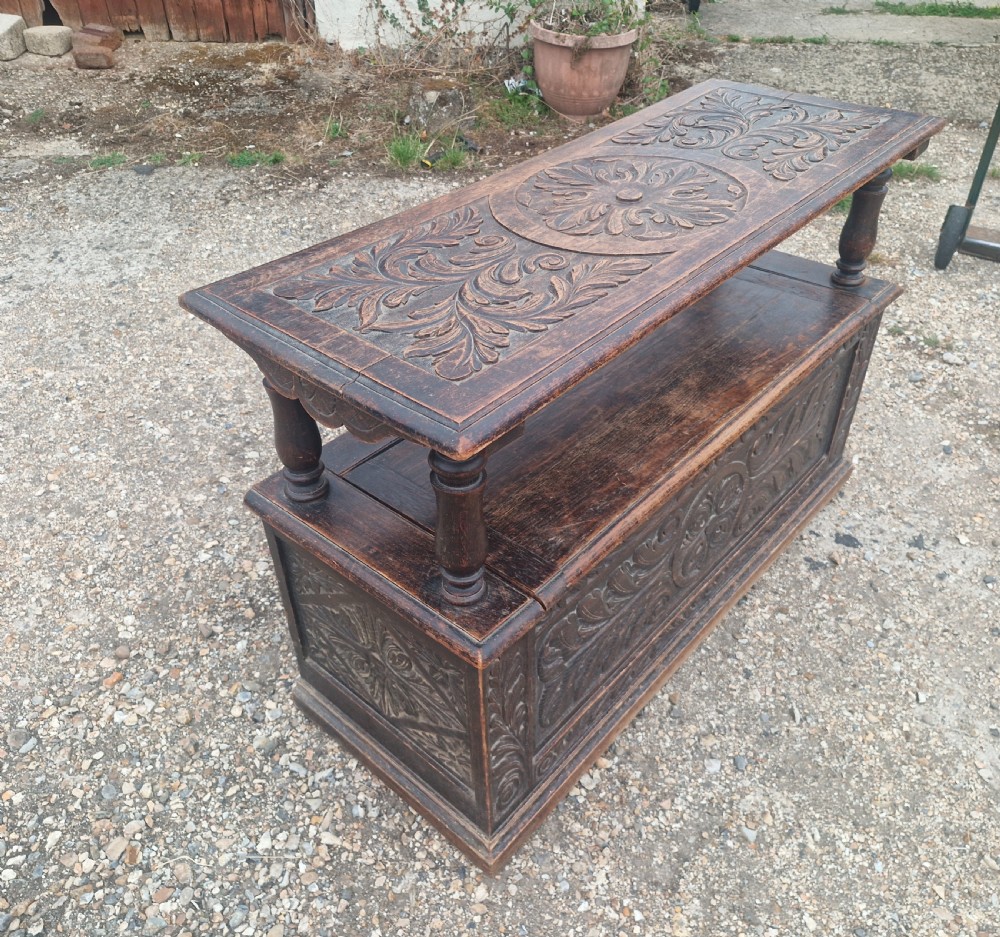 Late Victorian dark oak monks hall seat featuring intricate carved foliate panels, turned arm supports, and an integrated storage chest base. View VII