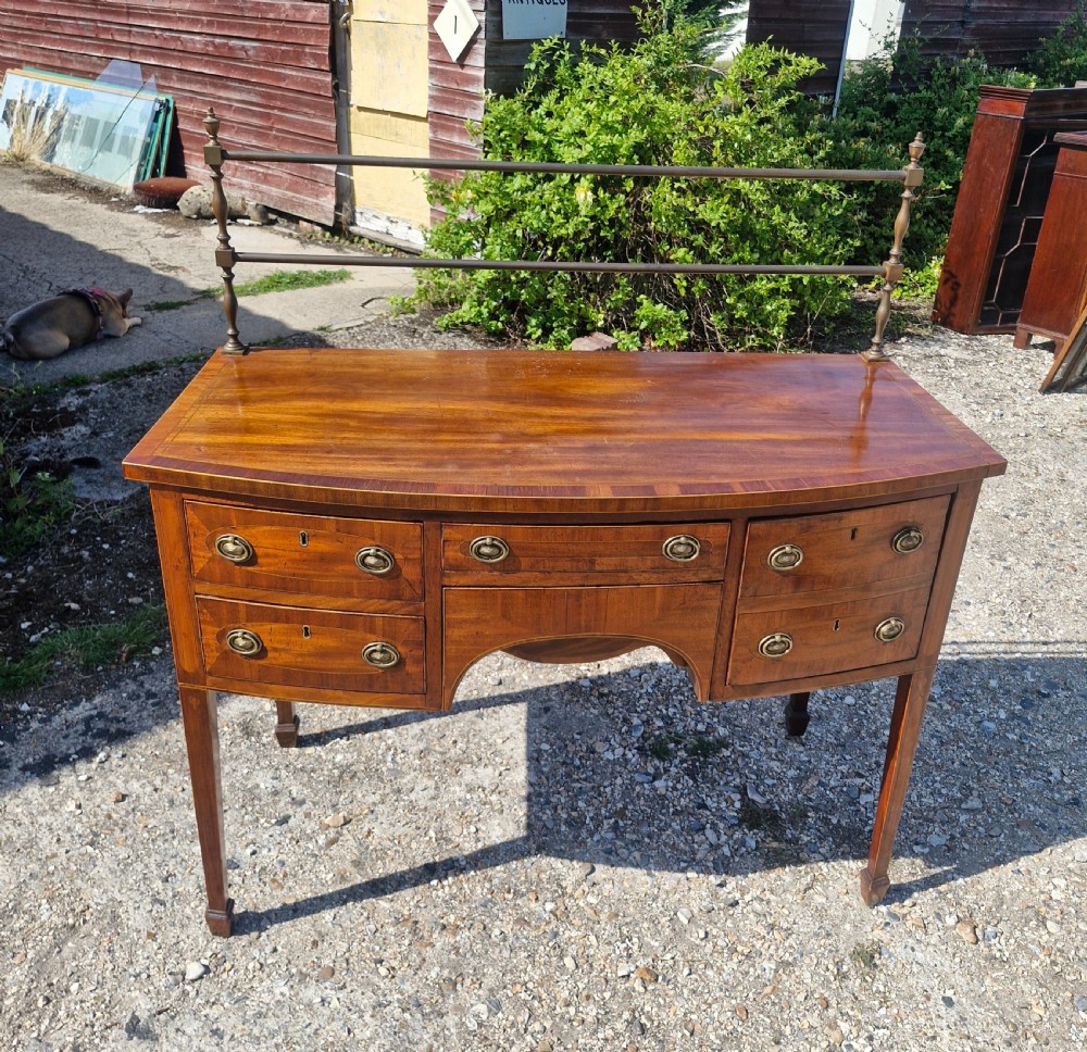 A late Georgian bowfront mahogany sideboard featuring boxwood inlay, brass ring handles, tapered legs, and a fitted brass gallery rail.