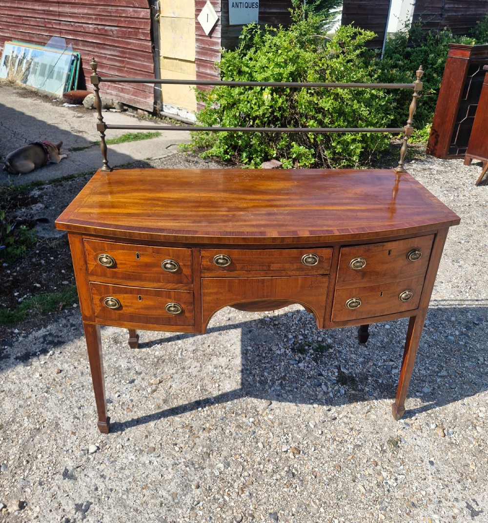 A late Georgian bowfront mahogany sideboard featuring boxwood inlay, brass ring handles, tapered legs, and a fitted brass gallery rail. View XI