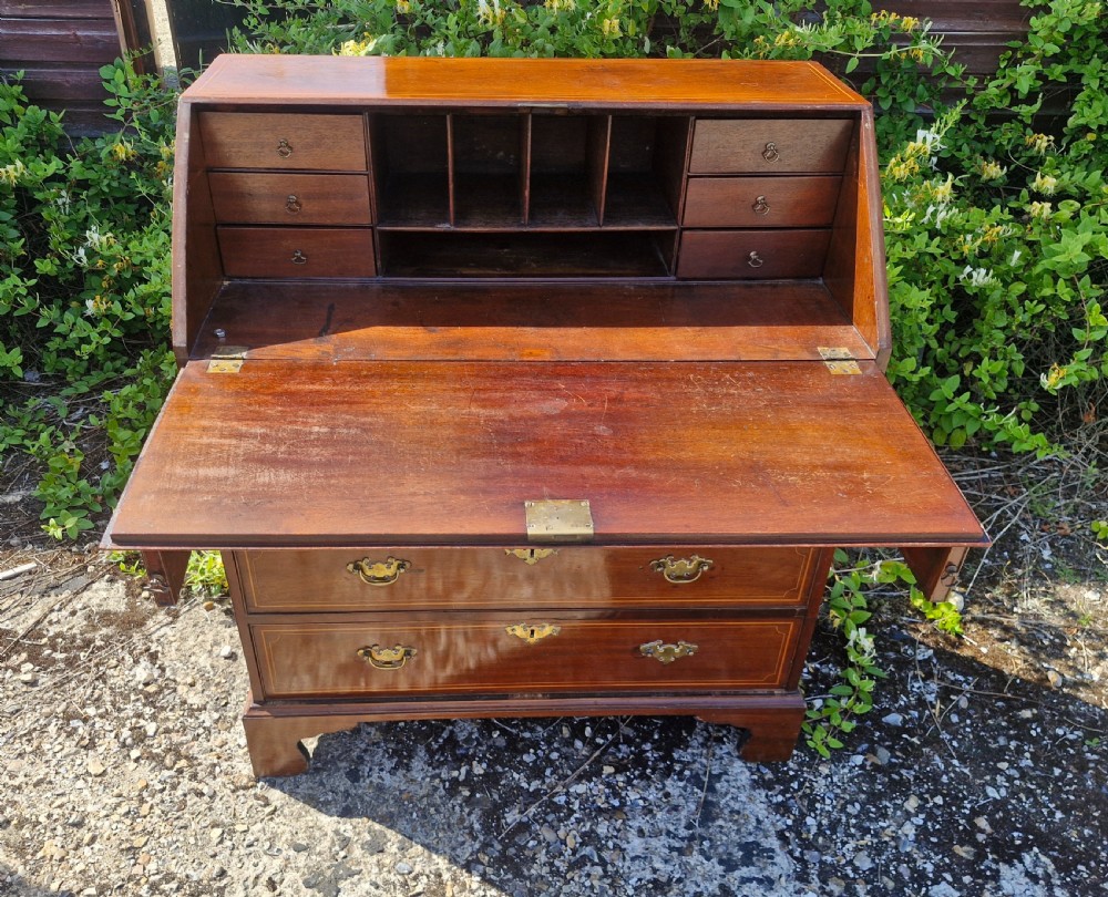 A late Georgian mahogany writing bureau featuring a sloped fall-front lid with boxwood inlay, four long drawers, brass swan-neck handles, and bracket feet. View V