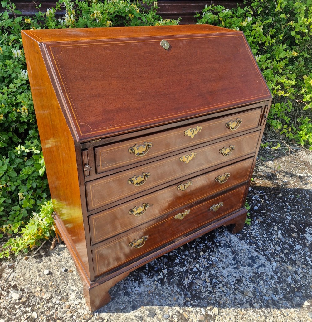 A late Georgian mahogany writing bureau featuring a sloped fall-front lid with boxwood inlay, four long drawers, brass swan-neck handles, and bracket feet. View IX