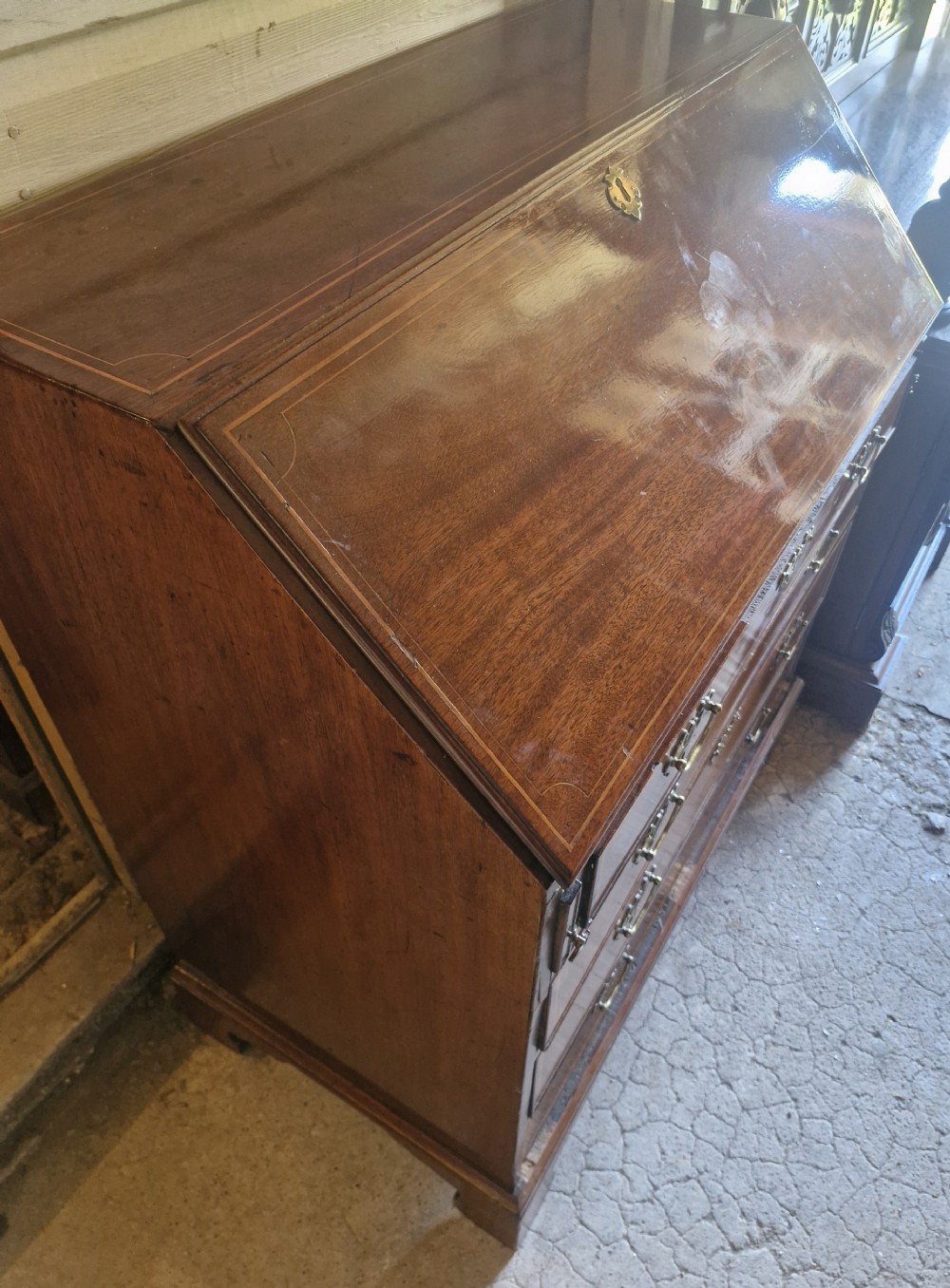A late Georgian mahogany writing bureau featuring a sloped fall-front lid with boxwood inlay, four long drawers, brass swan-neck handles, and bracket feet. View X