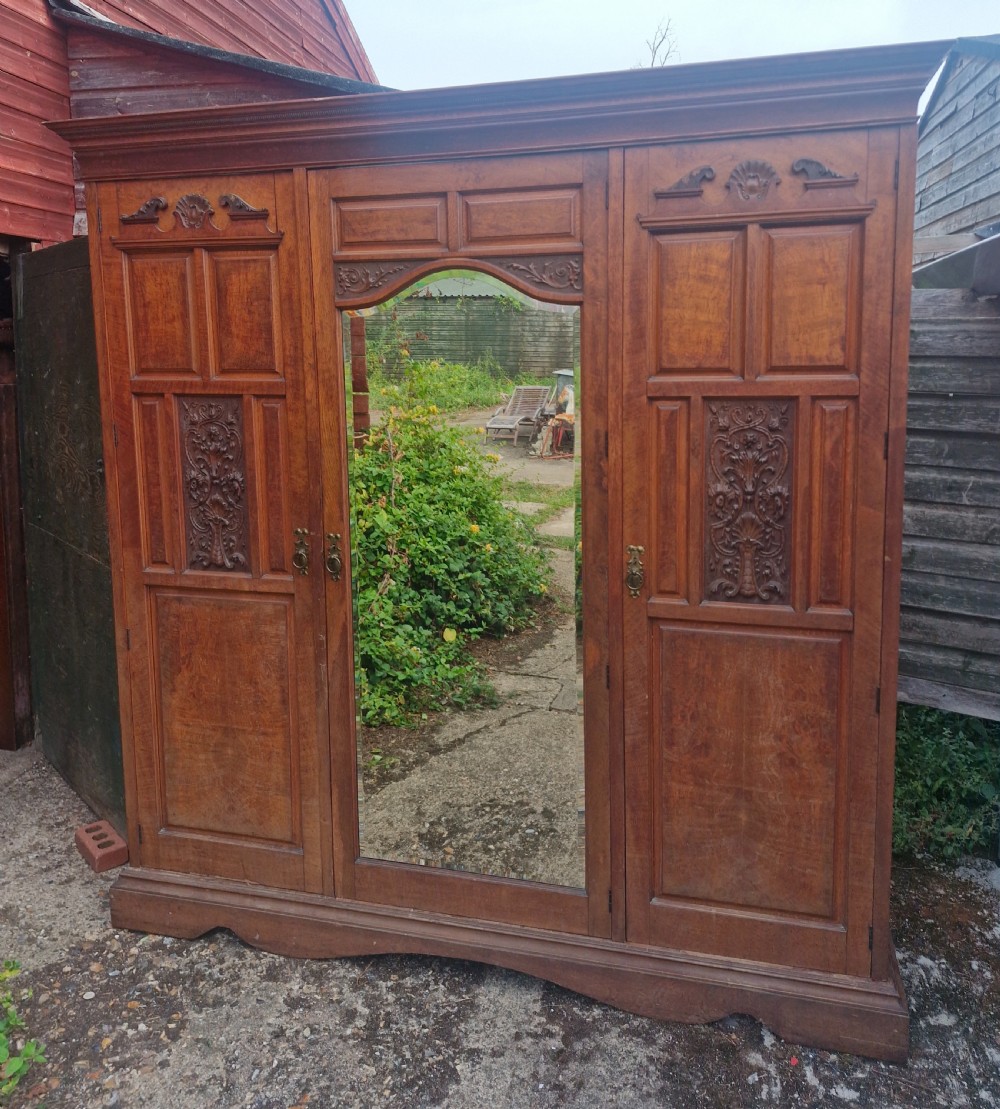 A large Edwardian oak triple wardrobe featuring carved side panels, an arched central mirror door, and traditional brass handles.
