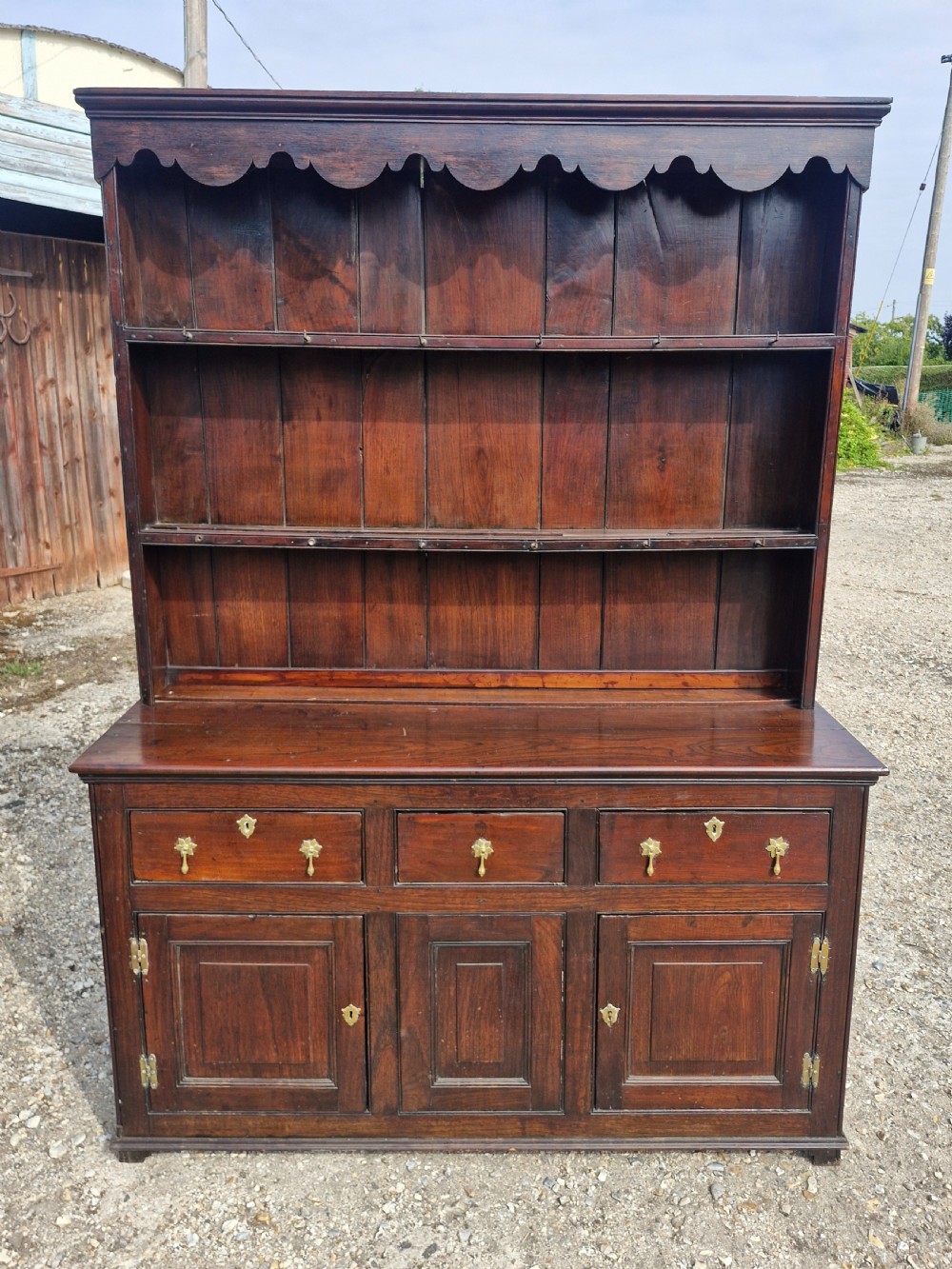 A nineteenth-century dark oak Welsh dresser featuring a scalloped upper plate rack above three drawers and three panelled cupboard doors with brass handles.