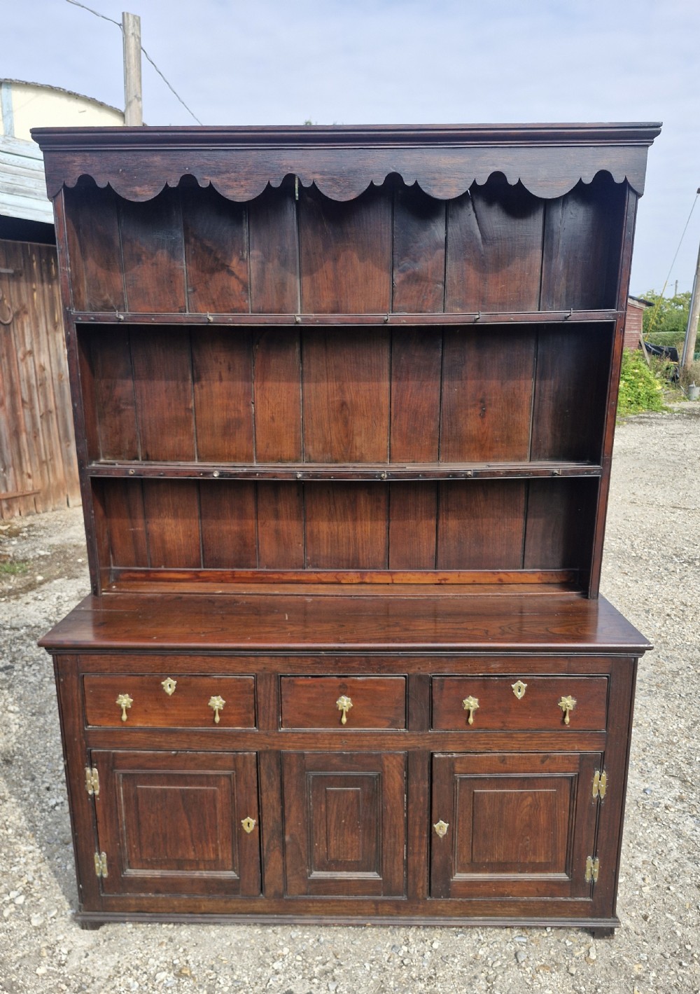 A nineteenth-century dark oak Welsh dresser featuring a scalloped upper plate rack above three drawers and three panelled cupboard doors with brass handles. View III