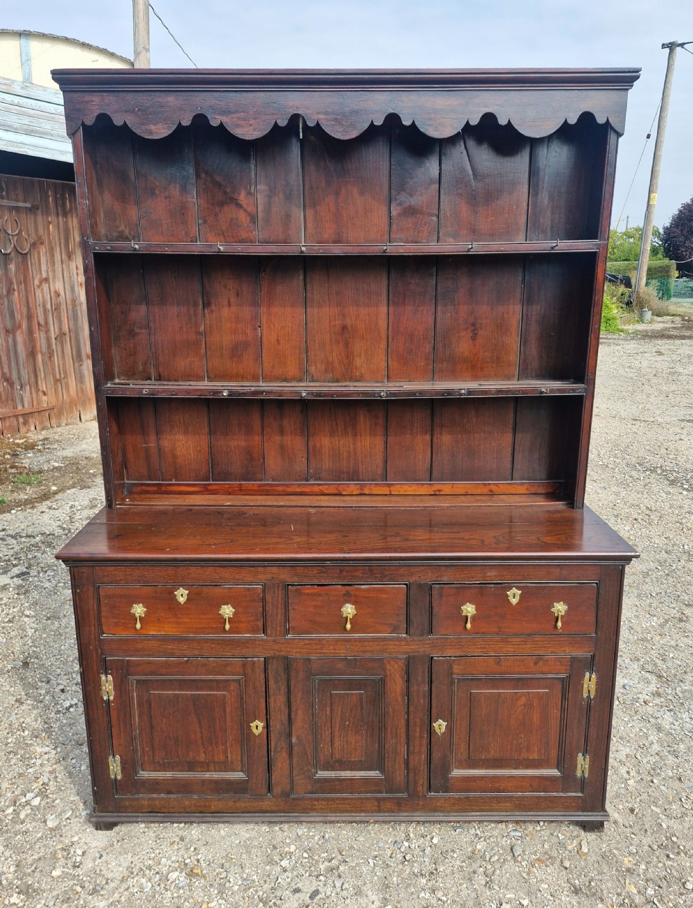 A nineteenth-century dark oak Welsh dresser featuring a scalloped upper plate rack above three drawers and three panelled cupboard doors with brass handles. View VII